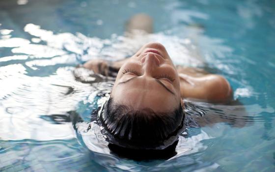Donna rilassata galleggia in piscina, occhi chiusi, acqua trasparente e calma.