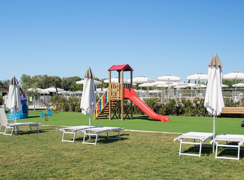 Area giochi all'aperto con scivolo, sdraio e ombrelloni su prato verde.